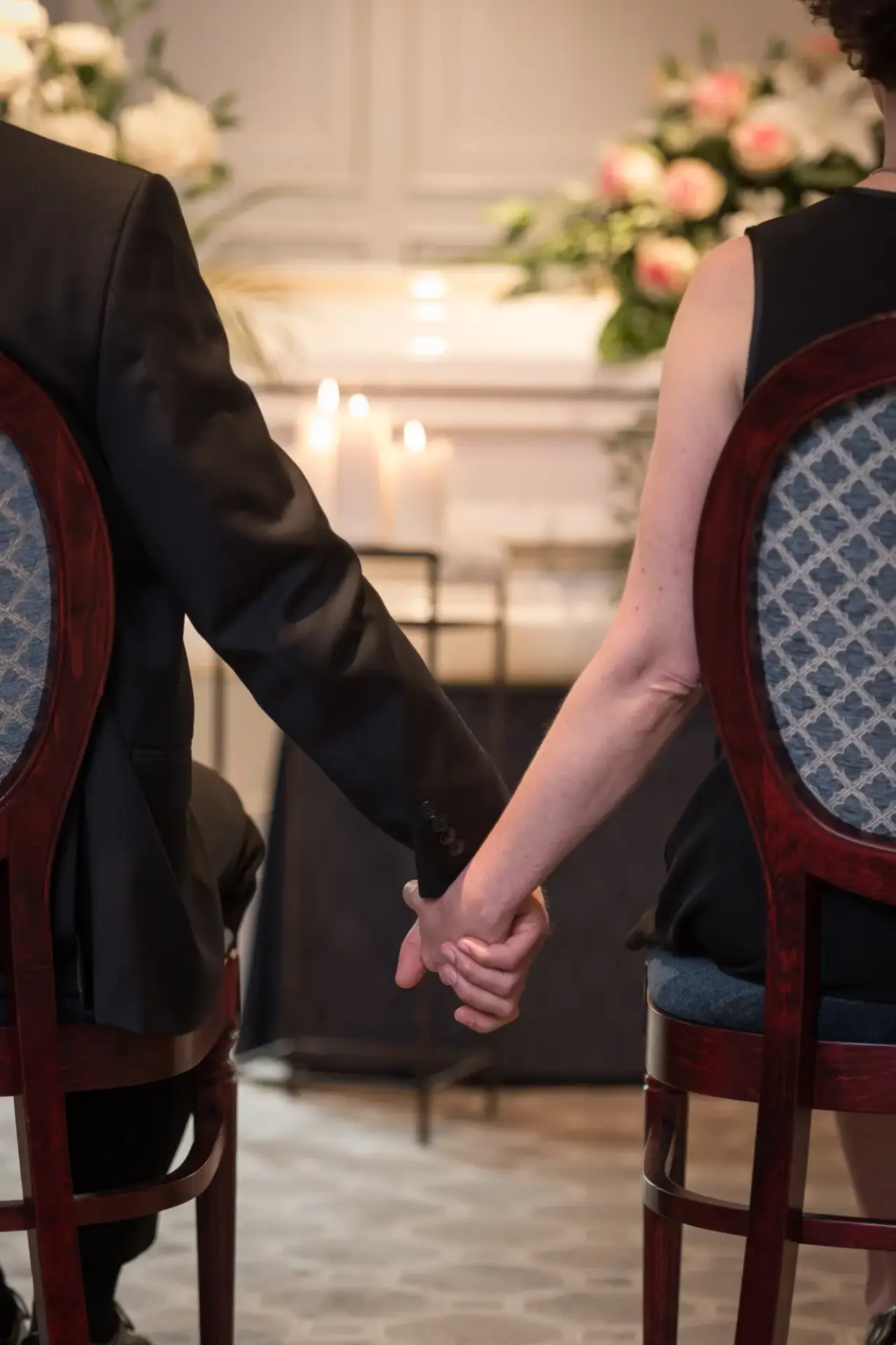 Two people holding hands while seated during a funeral service at Kane Fetterly, with flowers and candles in the background.
